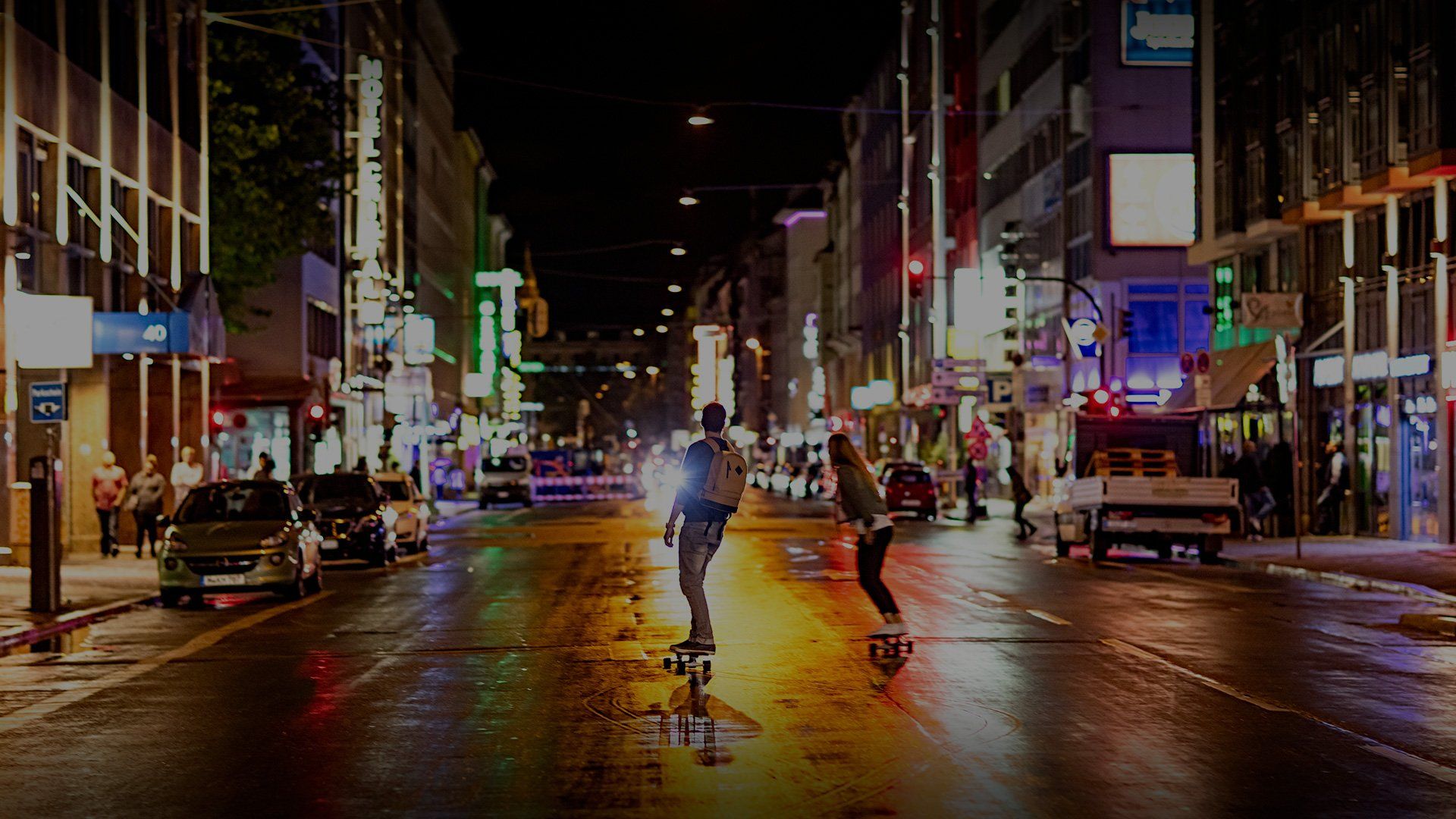 Two people skateboard down a city street at night, lights from buildings reflecting in the wet road surface.